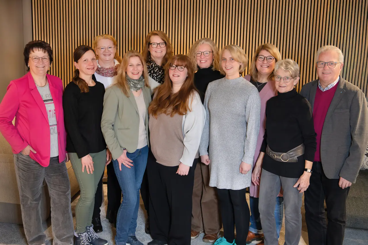 Elf Personen stehen in einer Reihe vor einer hellen Lamellenwand, tragen gemischte Kleidung in gedeckten und kräftigen Farben und stehen dicht beieinander.